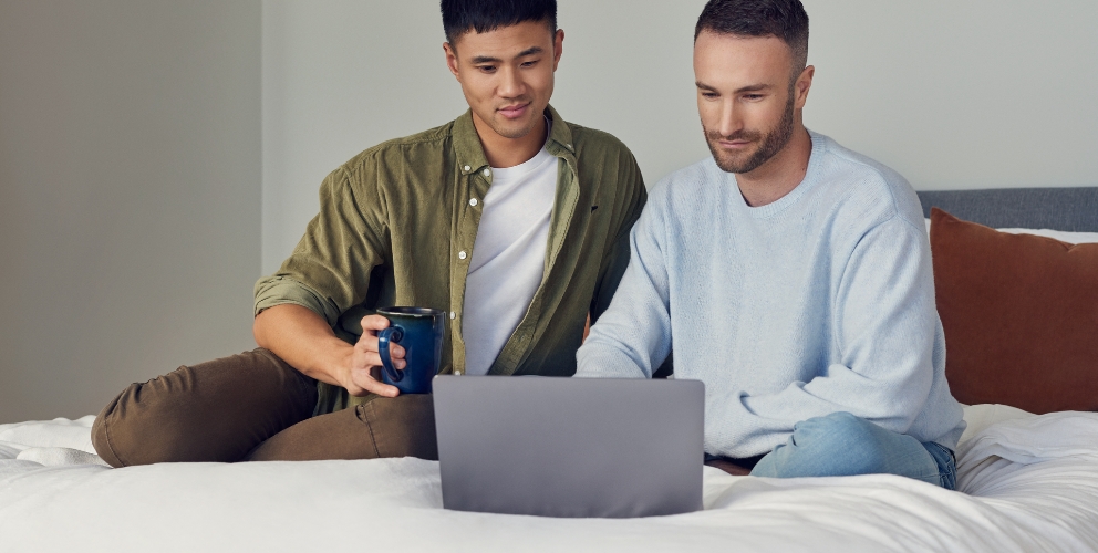Zwei Männer sitzen entspannt auf einem Bett und schauen auf einen Laptop.